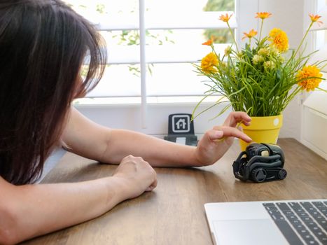 A woman petting a small robot on a wooden table beside a laptop and flowers.