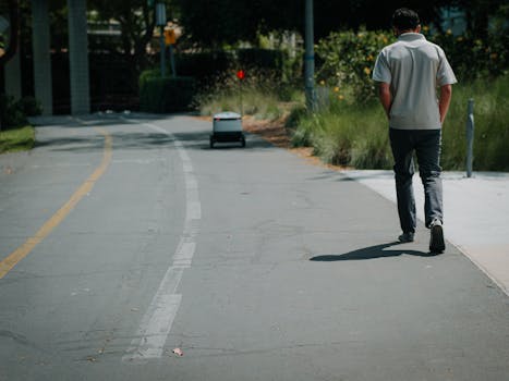 A man walks down a sidewalk beside a small delivery robot on a sunny day.
