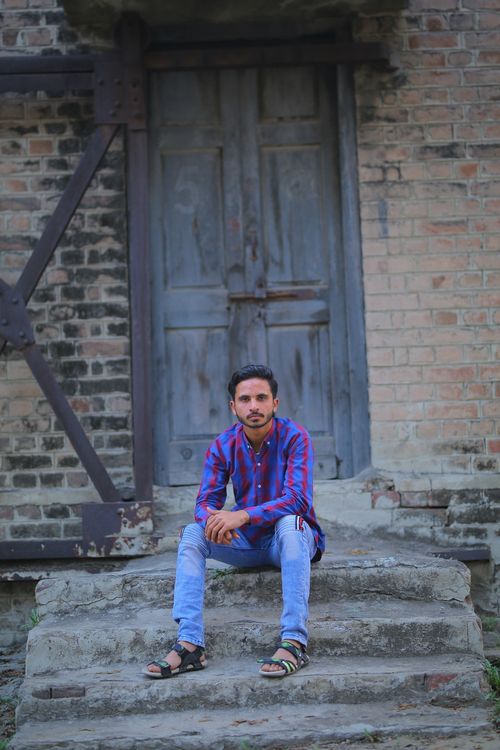Free A man with short hair and casual attire sitting on steps in front of a rustic blue wooden door. Stock Photo