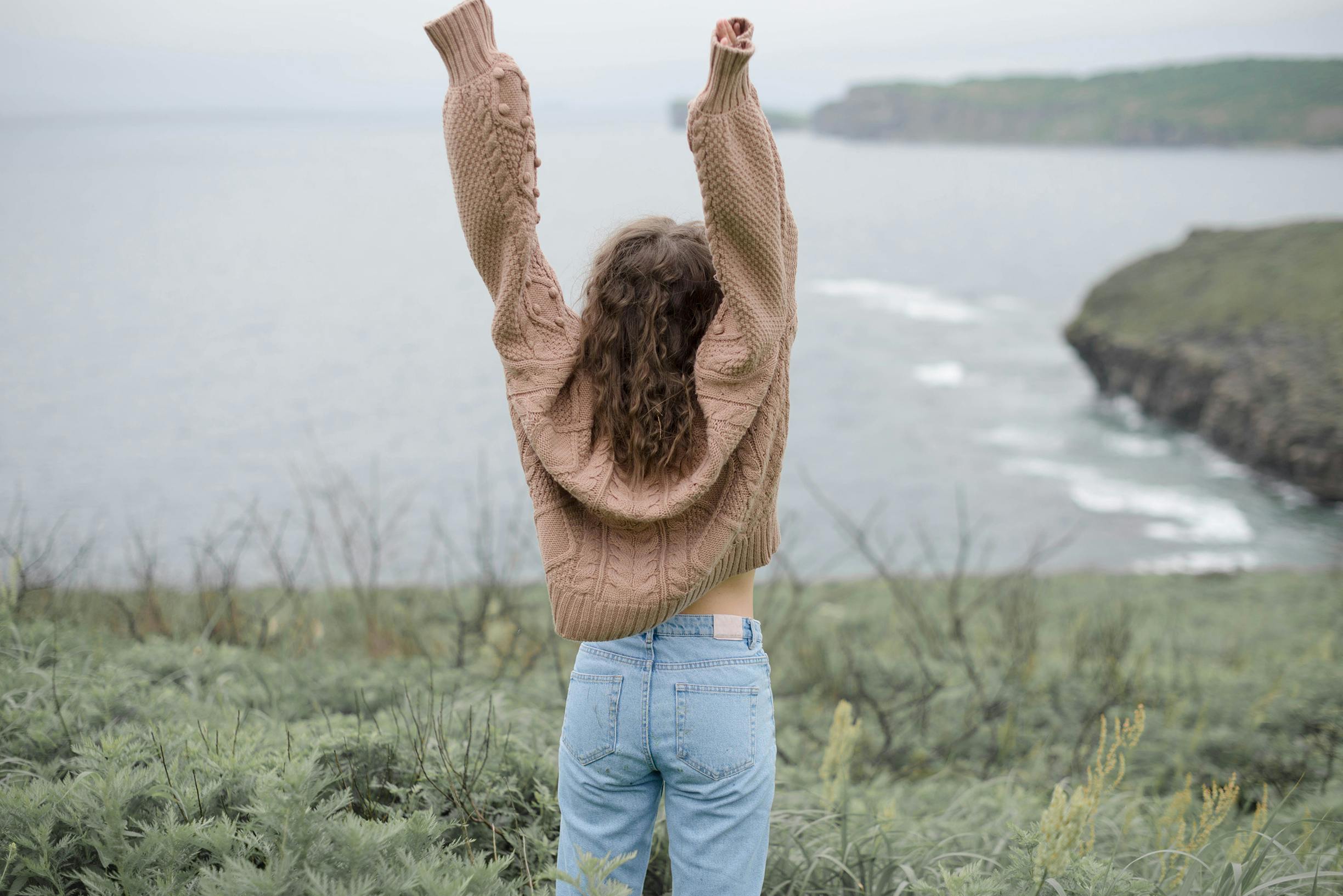 一名穿着针织毛衣和牛仔裤的女子在海边高兴地举起双臂