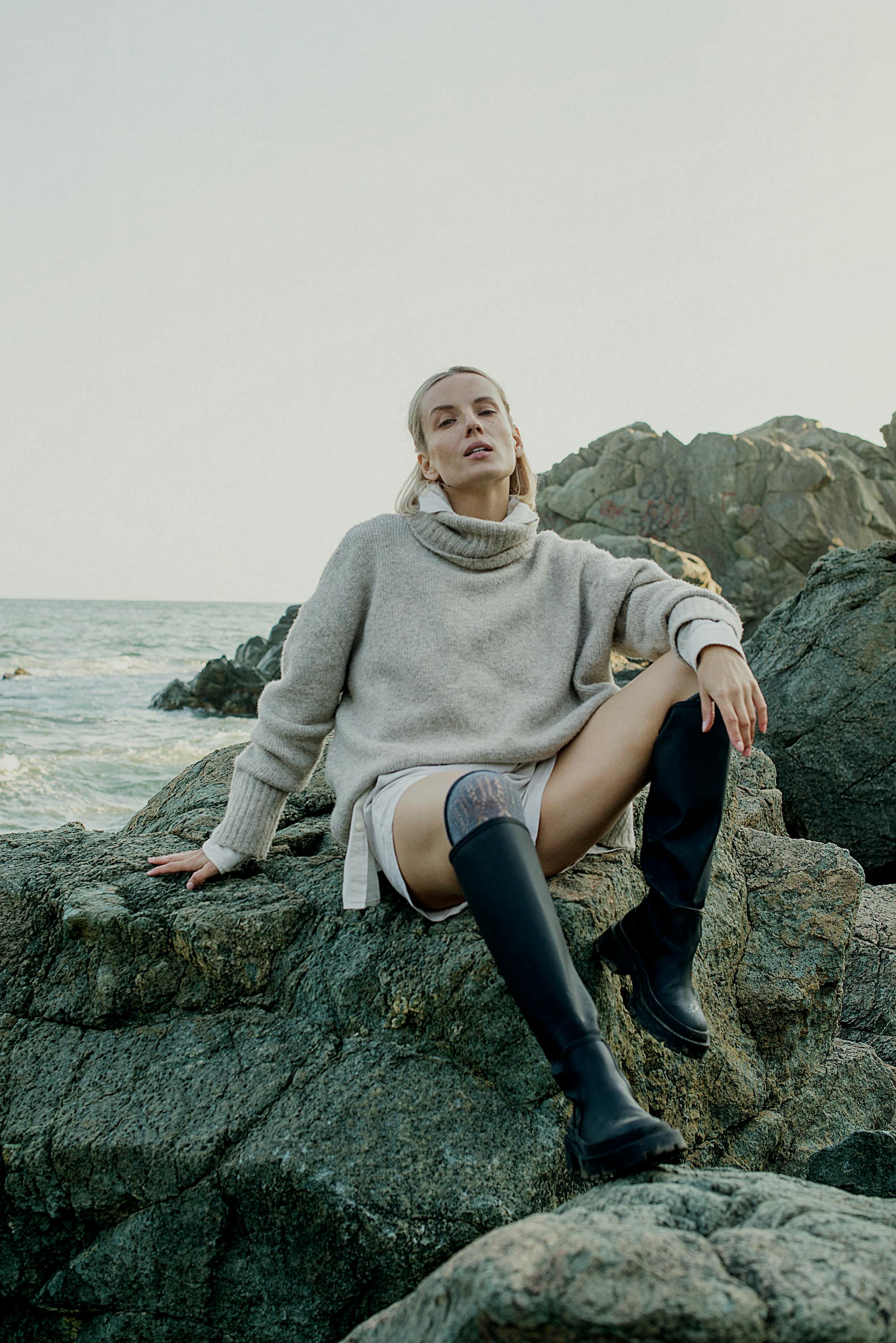 晴朗的日子里，一名穿着舒适毛衣和靴子的女子在海边的岩石上摆姿势