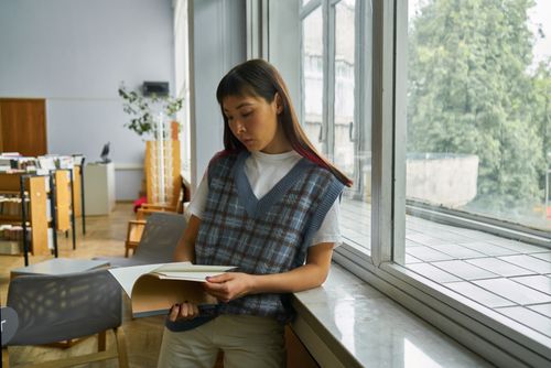 Free A young woman in a library, leaning on a window sill, deeply immersed in a book. Stock Photo