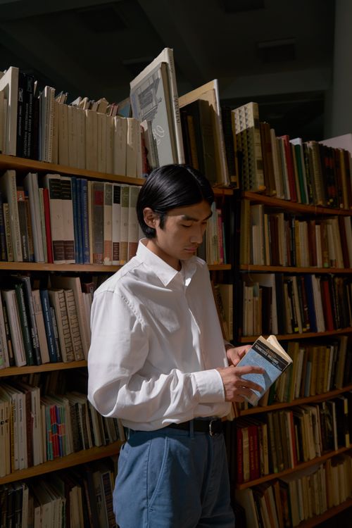 Free Young Asian man reading a book in a library, standing among bookshelves. Stock Photo