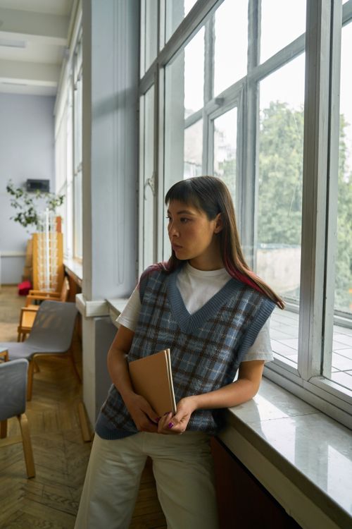 Free A stylish woman stands by a large window, holding a notebook and gazing thoughtfully away. Stock Photo