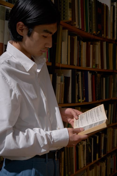 Free Young man in a library reading a book, surrounded by shelves of books. Ideal for educational themes. Stock Photo
