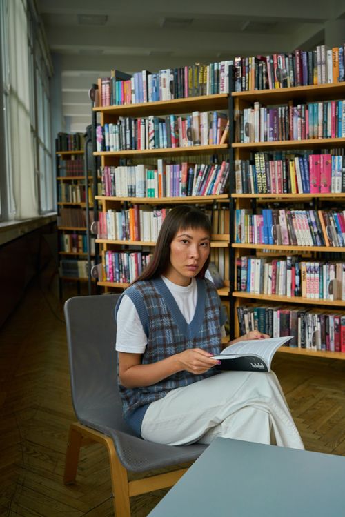 Free A woman sitting comfortably while reading a book in a well-lit library with shelves full of books. Stock Photo