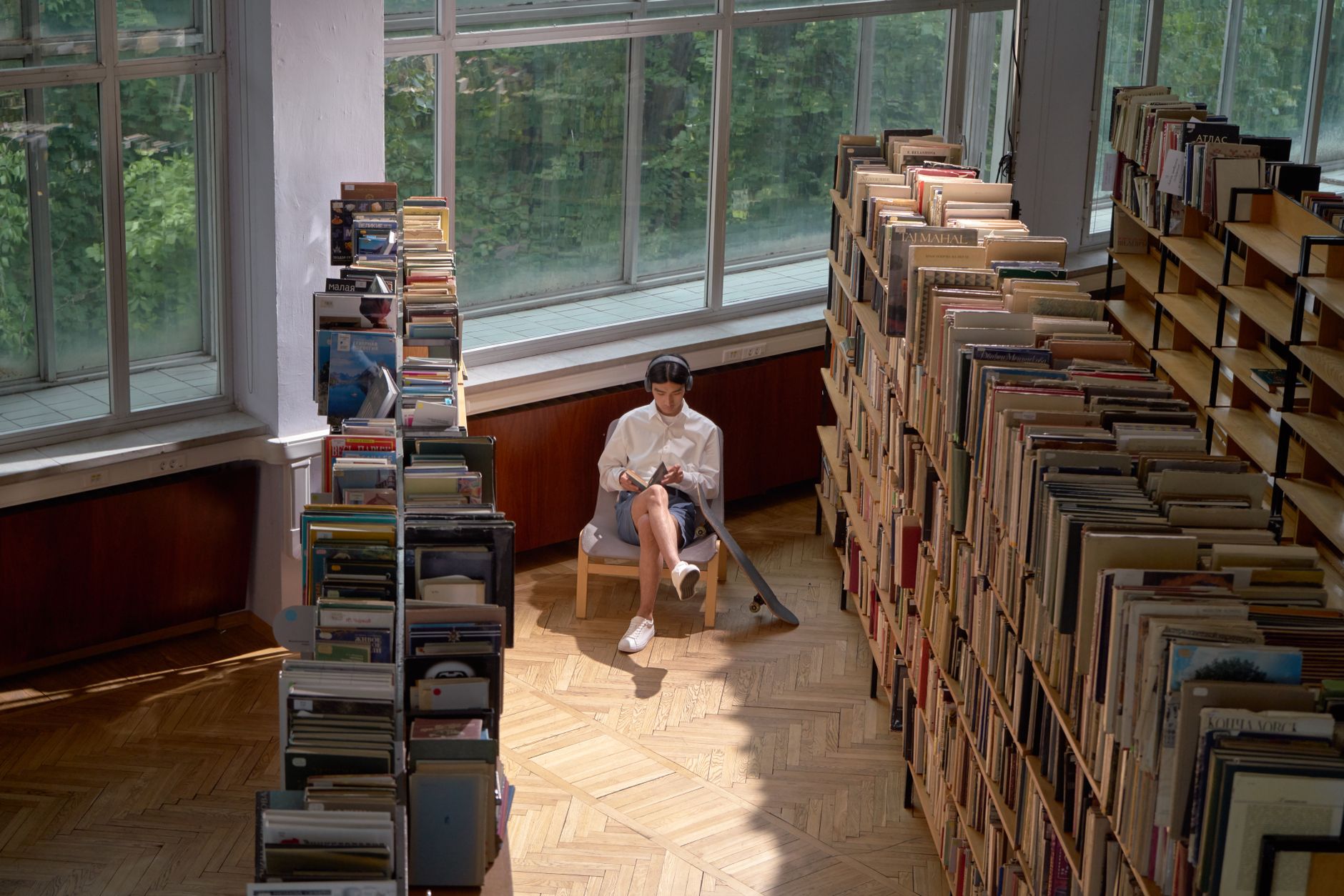 A young adult reads a book in a quiet, sunlit library surrounded by shelves of books.