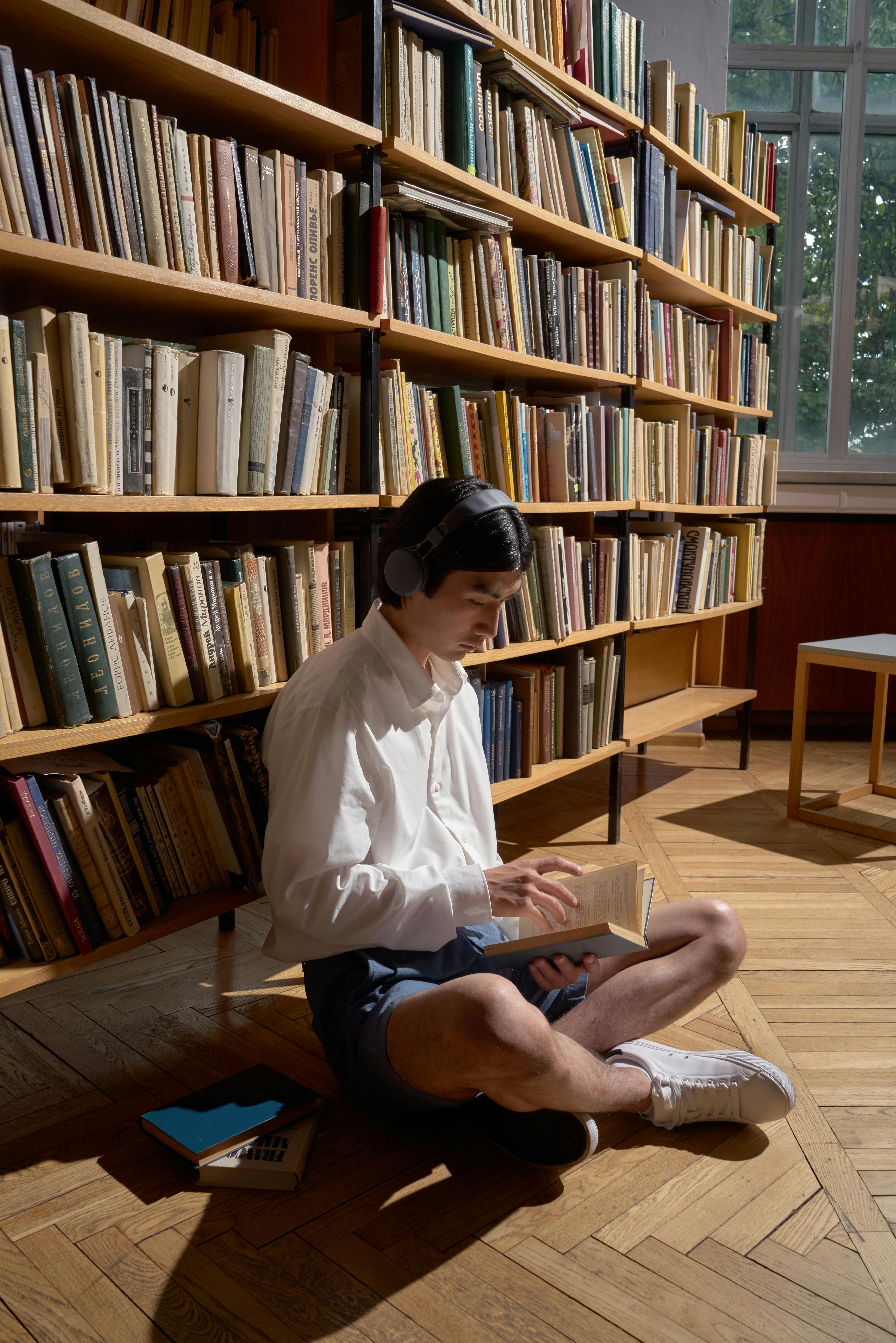 Free Young Asian man sitting in library reading a book with headphones on. Stock Photo