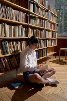 Young Asian man sitting in library reading a book with headphones on.