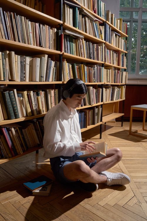 Free Young Asian man sitting in library reading a book with headphones on. Stock Photo