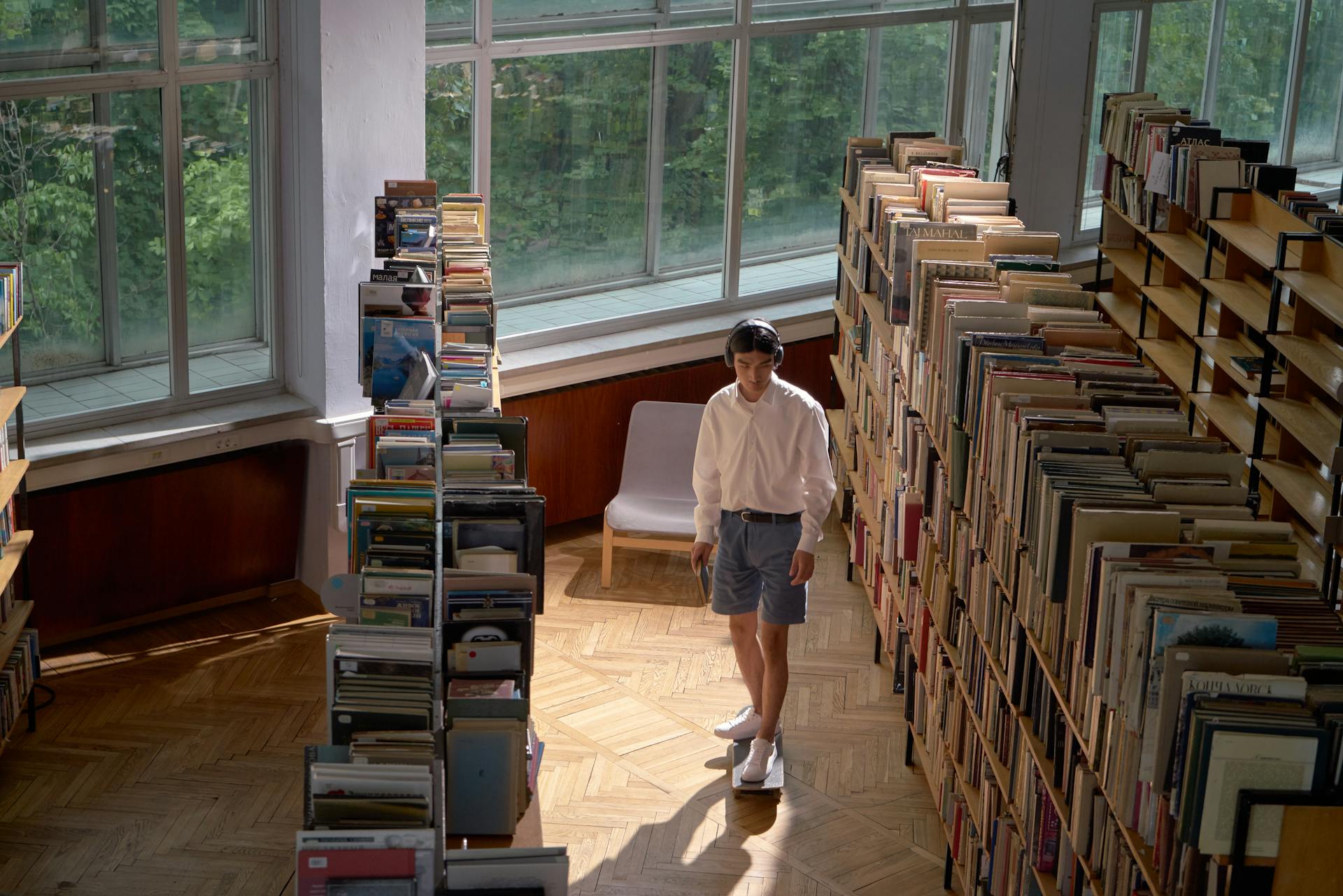 Asian man with headphones in a library aisle enjoying music under sunlight.