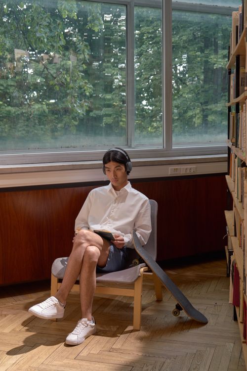 Free Asian man sitting in a library, wearing headphones, reading a book with a skateboard leaning against the chair. Stock Photo