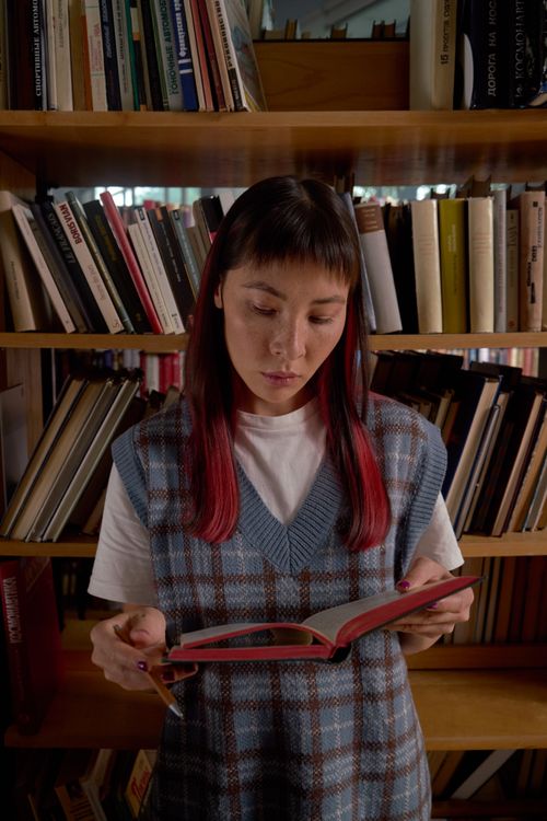 Free Asian woman with colored hair reading a book in a library, wearing a sweater vest. Stock Photo