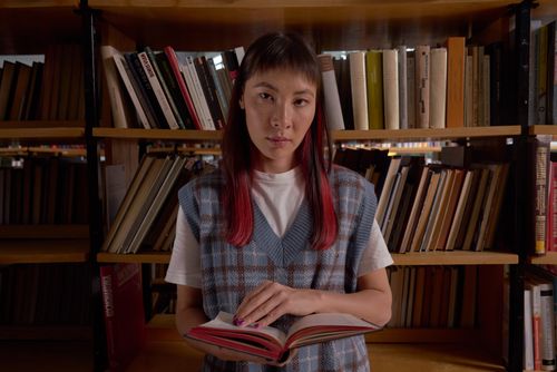 Free A young woman stands in a library holding an open book, surrounded by bookshelves. Stock Photo