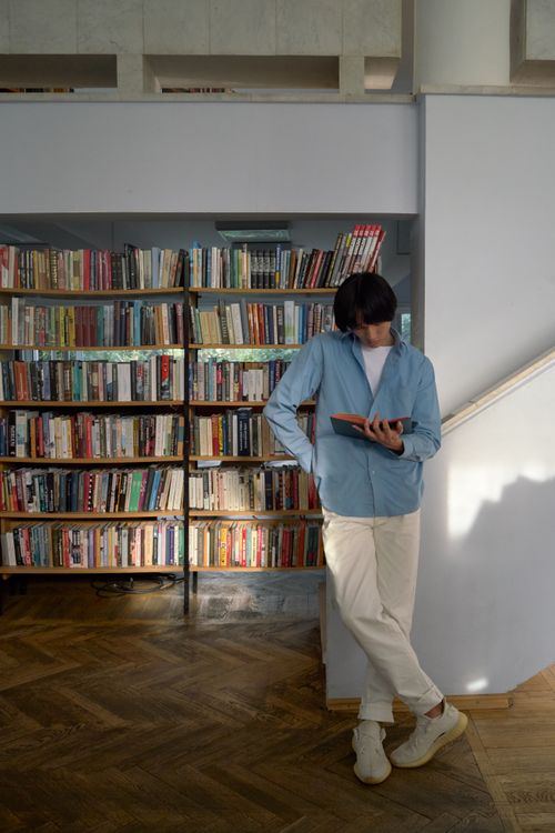 Free A young man in casual attire leans against a library wall, immersed in reading. Stock Photo