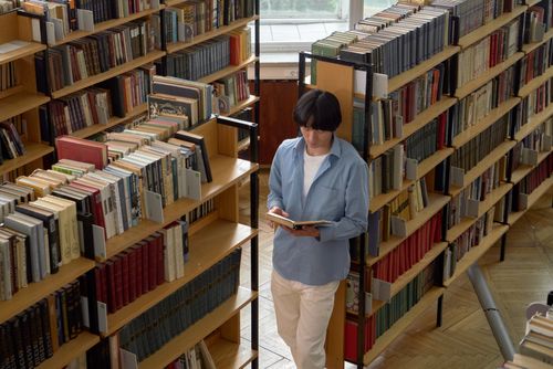 Free A young man reads intently in a bright library surrounded by bookshelves. Stock Photo
