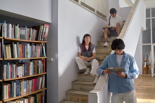 Free Three students reading on a staircase near bookshelves inside a library setting. Stock Photo