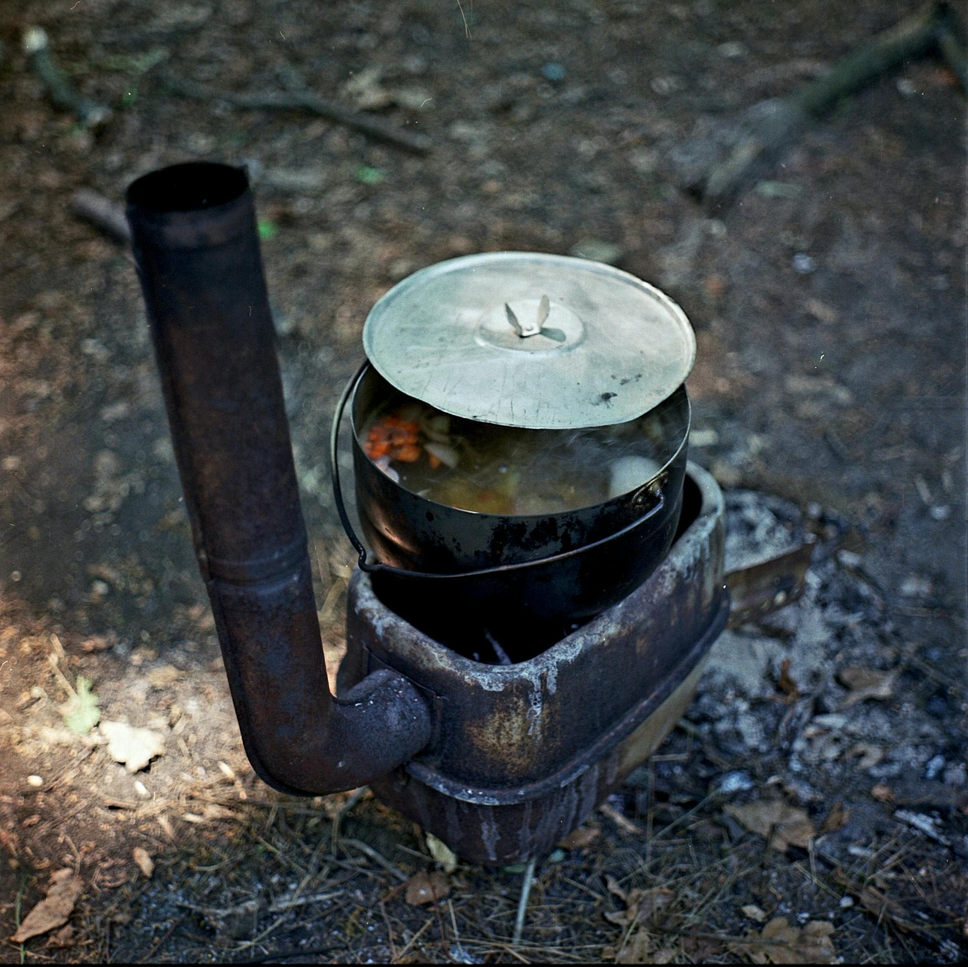 A rustic wood stove with a pot of food outdoors in a natural setting.