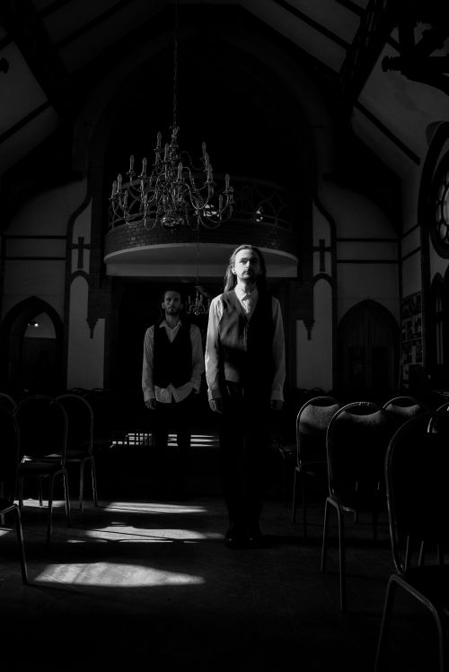 Free Two men in vests walk through a shadowed church interior under a chandelier. Stock Photo