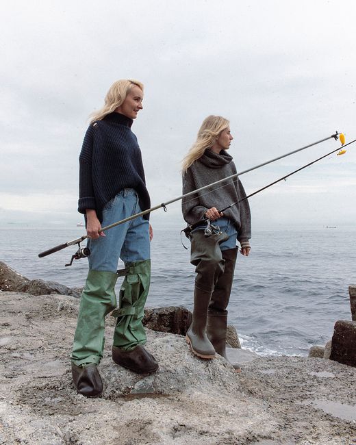 Two women fishing on a rocky seaside cliff in daytime, wearing rubber boots and sweaters.