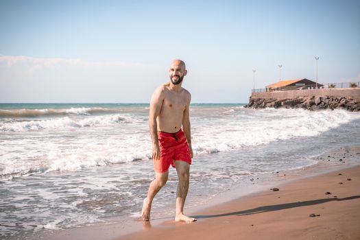 A bald man walks shirtless on İskenderun beach, enjoying the summer waves.