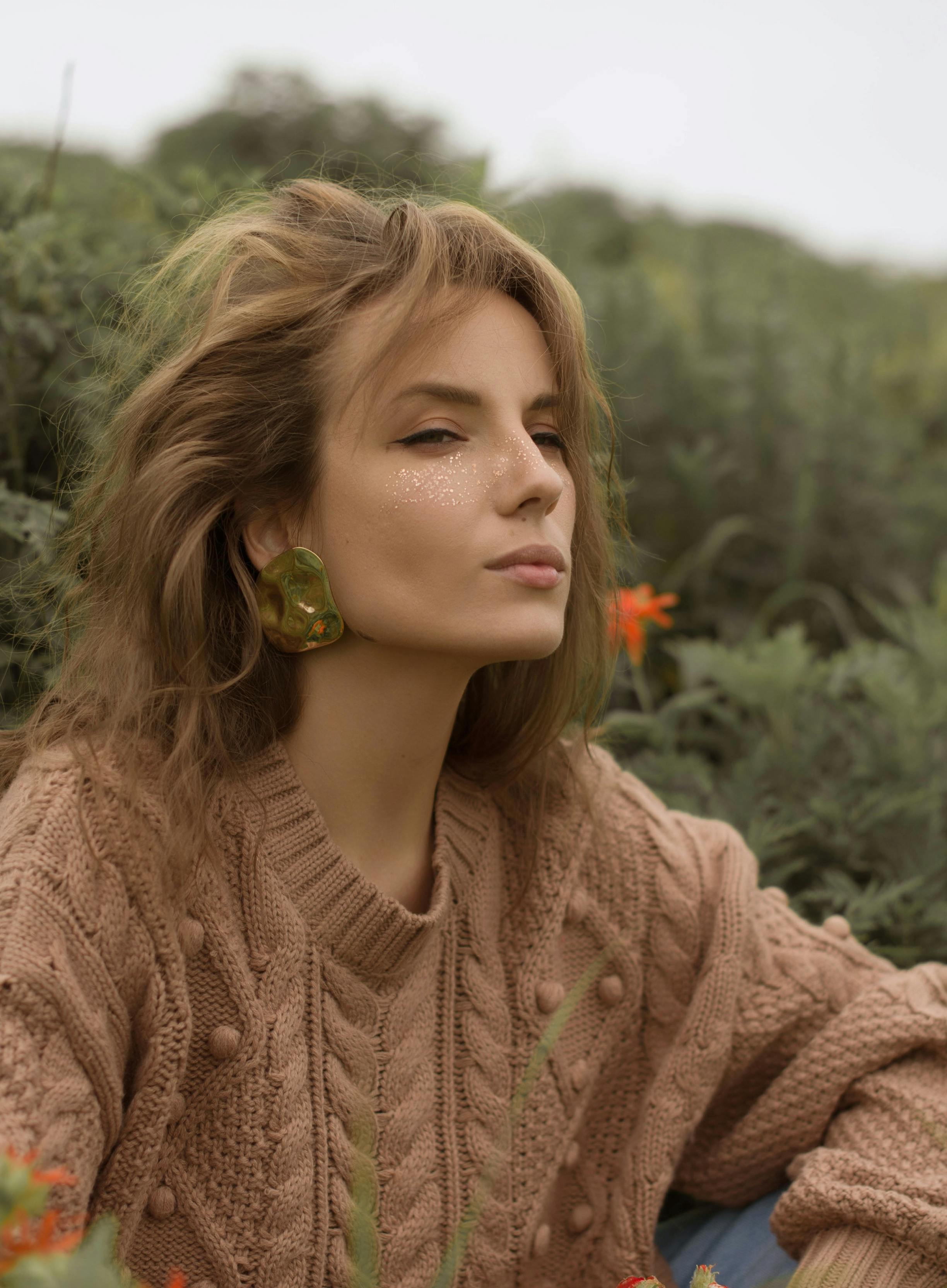 Fashionable woman in brown sweater with makeup, posing amidst greenery, evoking earthy tones.