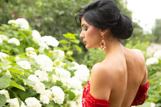 A woman in a glamorous red dress stands gracefully among blooming hydrangeas in an outdoor setting.