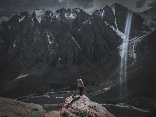 Free Man stands on rocky terrain, gazing at towering mountains with dramatic light beam. Stock Photo