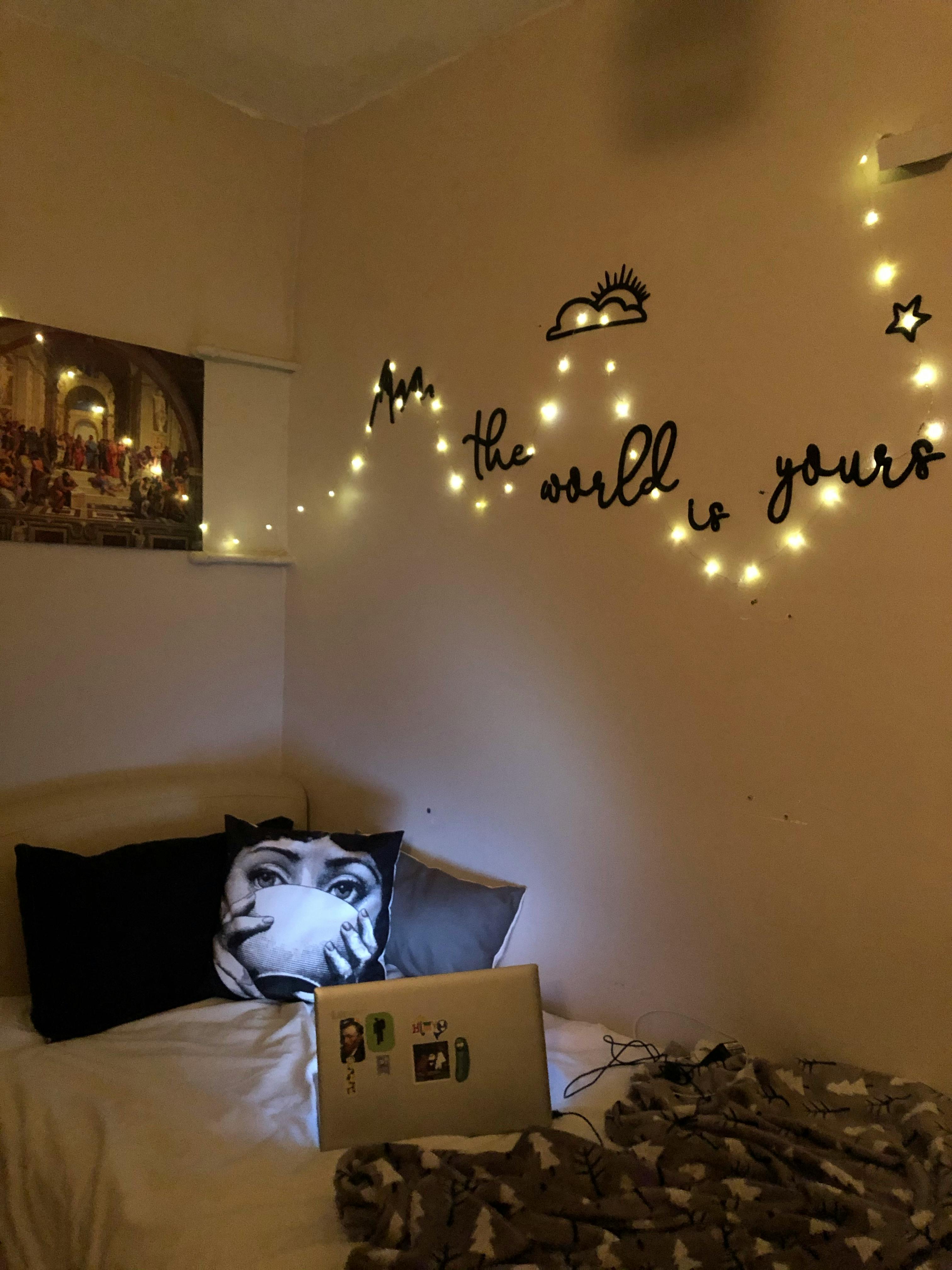 Free A cozy bedroom with string lights, a pillow, and a laptop on the bed, creating a warm ambiance. Stock Photo