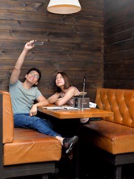 Asian couple sitting in a cozy cafe booth taking a selfie with a mobile phone.