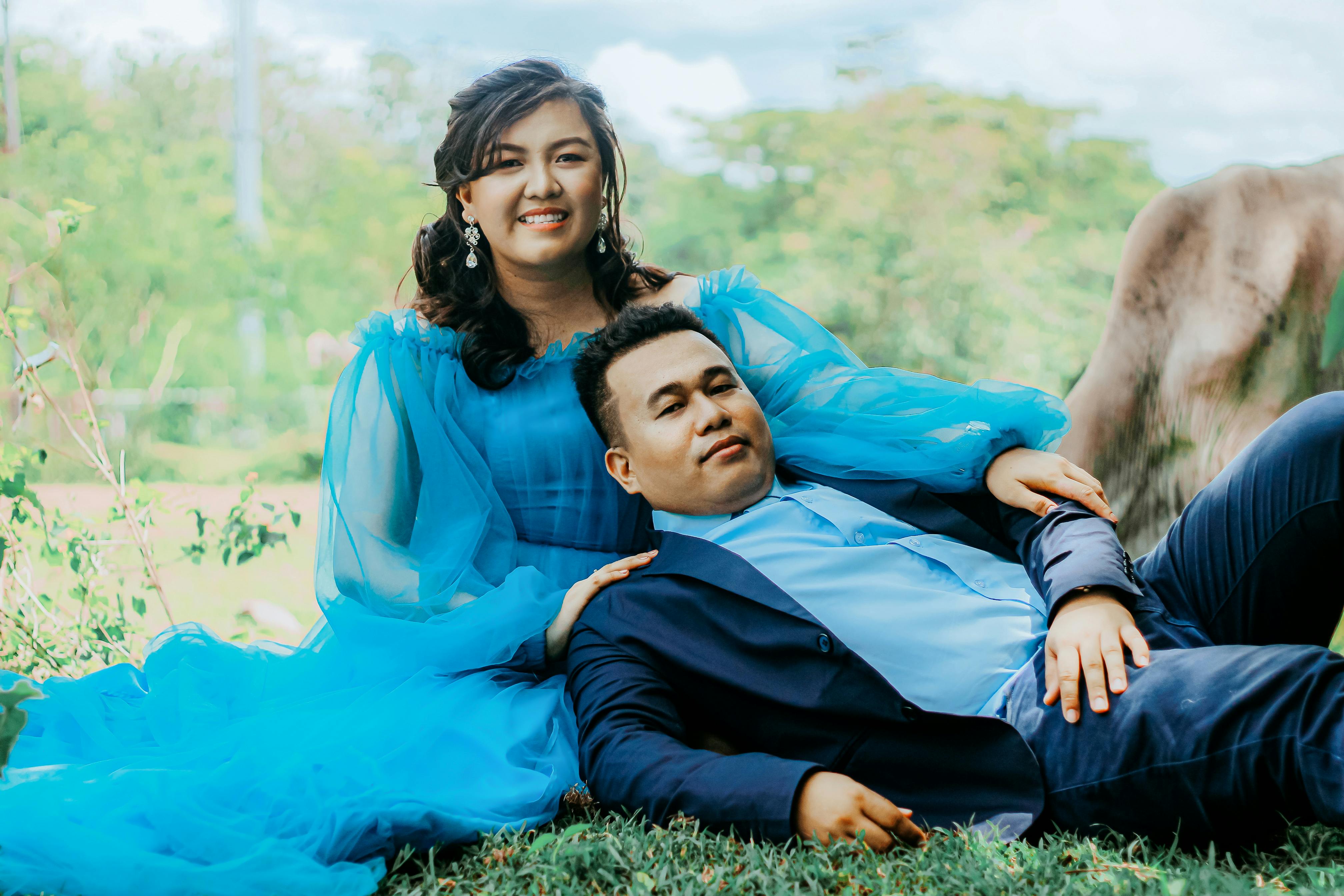 An elegant couple in blue attire enjoys a relaxing moment outdoors, surrounded by greenery.