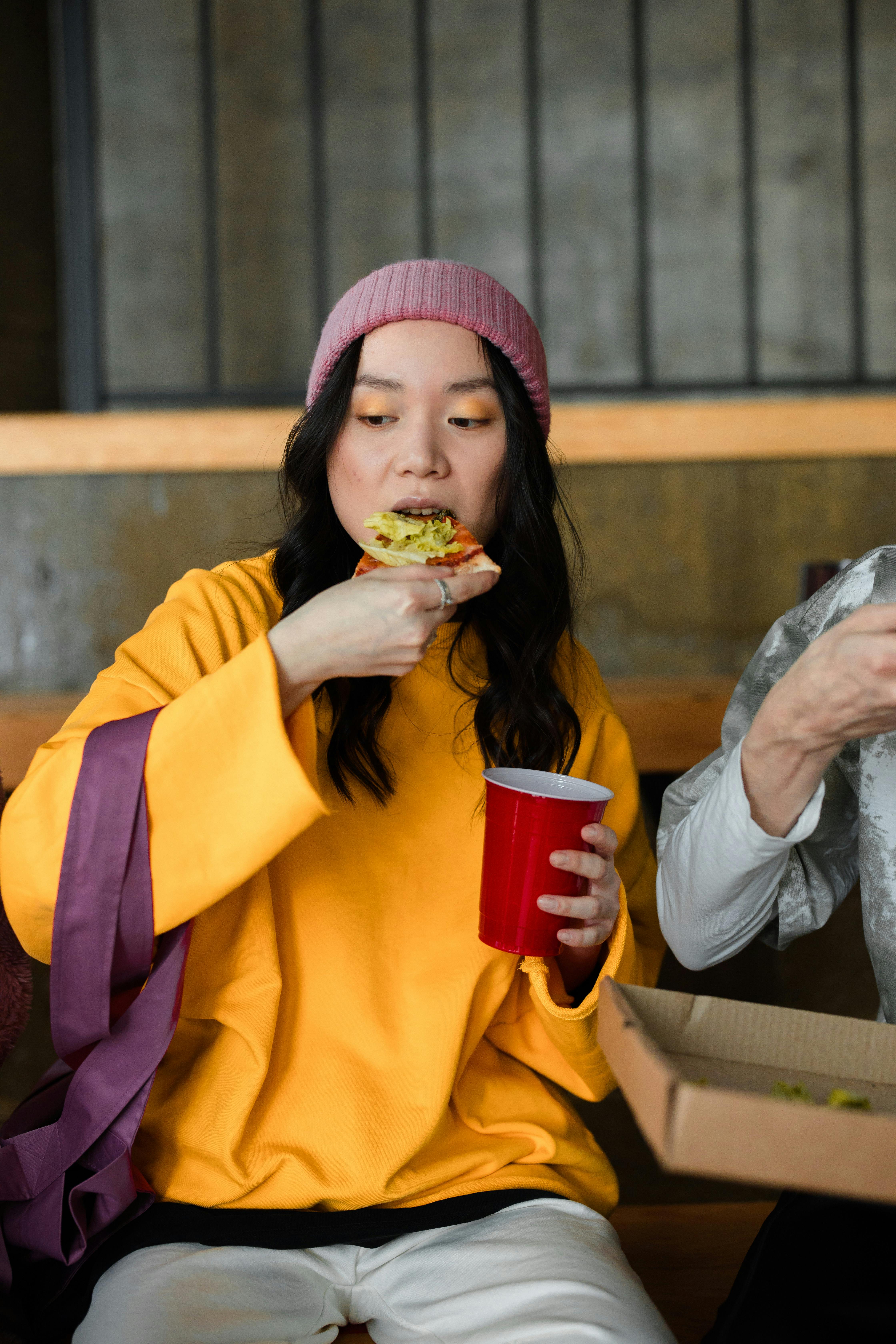 Asian woman in a beanie enjoying pizza with a red cup indoors.