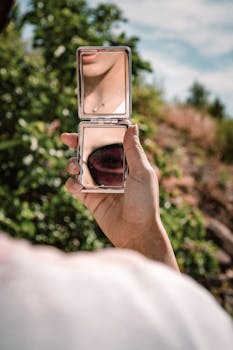 Close-up of a hand holding a pocket mirror showing a reflection outdoors in summer.