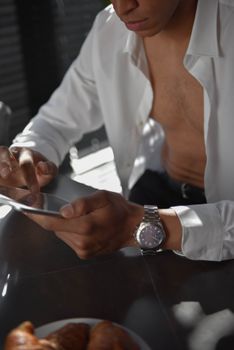 A stylish young man in a white shirt uses a digital tablet while enjoying breakfast in natural light.