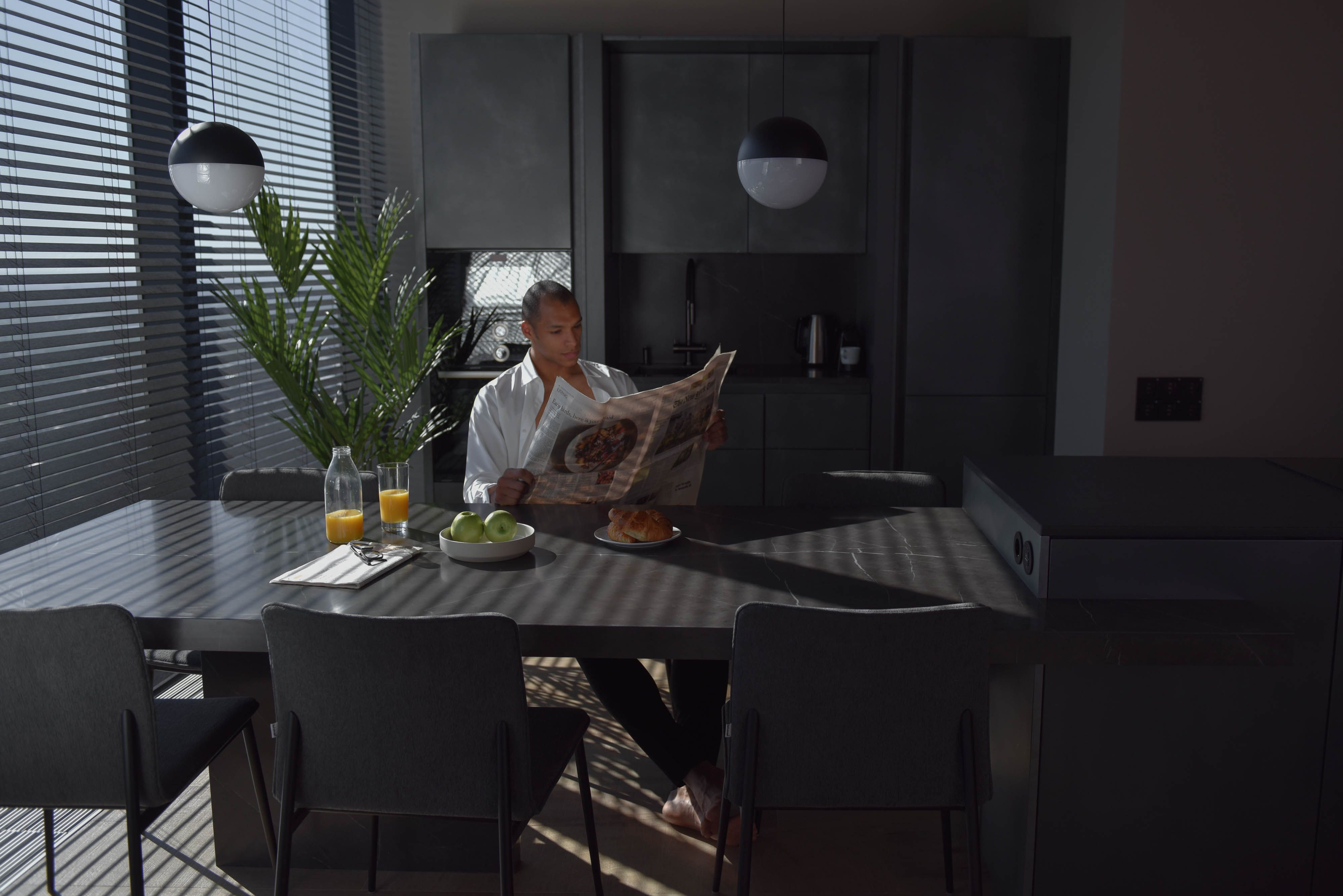 A man reads a newspaper at a modern dining table with green apples and orange juice.