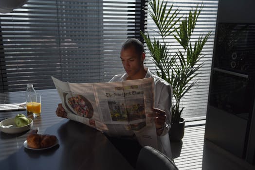 Adult reading newspaper with breakfast in modern kitchen, morning sunlight.