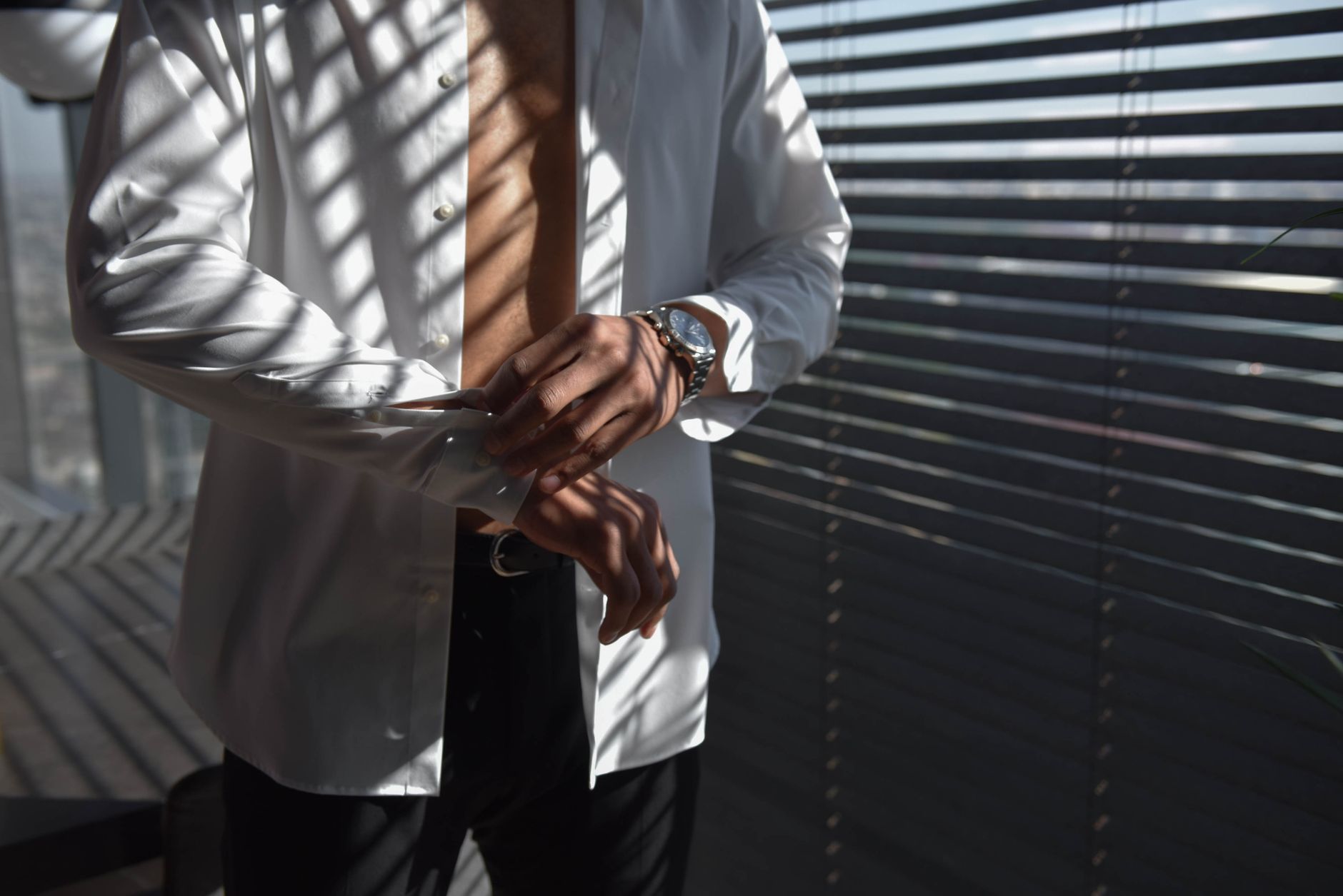 A fashionable man adjusting his shirt cuffs in sunlight filtered through blinds in a modern interior space.