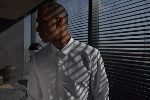 Free A stylish businessman in white shirt stands next to window blinds casting shadows. Stock Photo