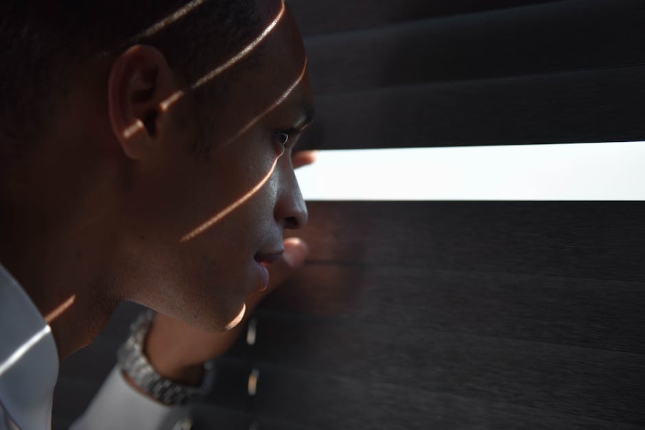 Profile view of a man curiously peeking through window blinds with shadows on his face.