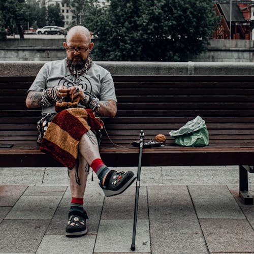 Bald Man in Eyeglasses with Bracelets Sitting on Wooden Bench While Knitting