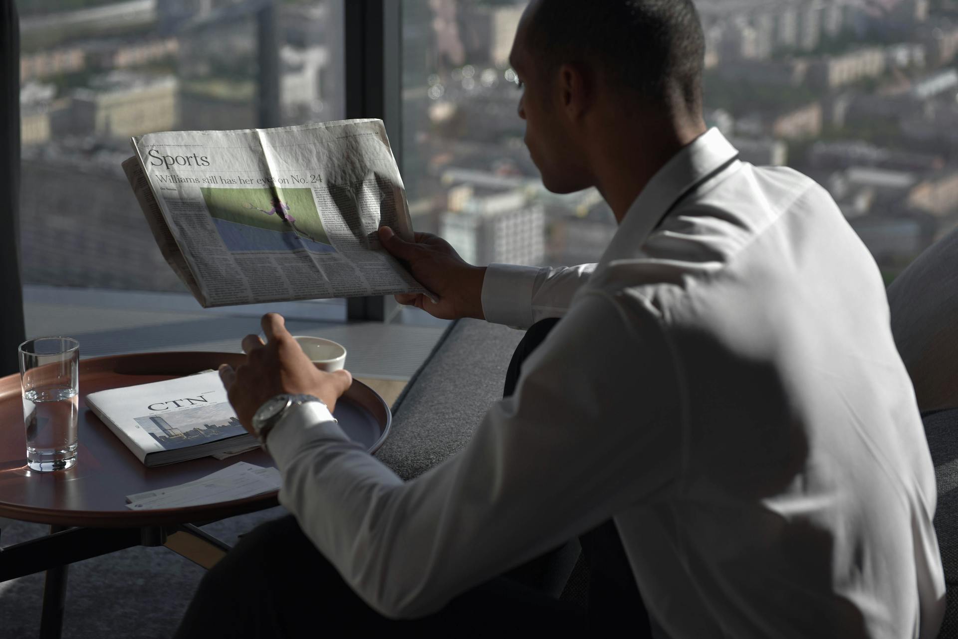 Businessman in office reading sports section, enjoying coffee with city view.