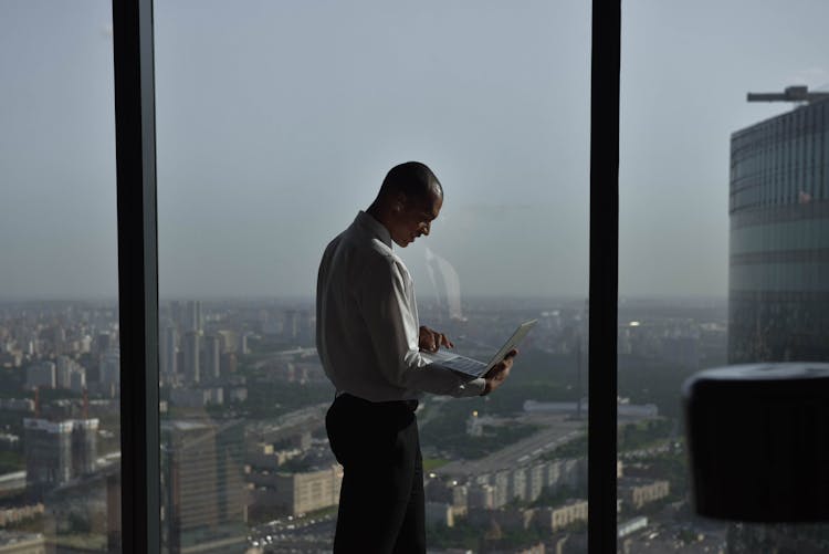 Man In Shirt Standing With Laptop By Windows