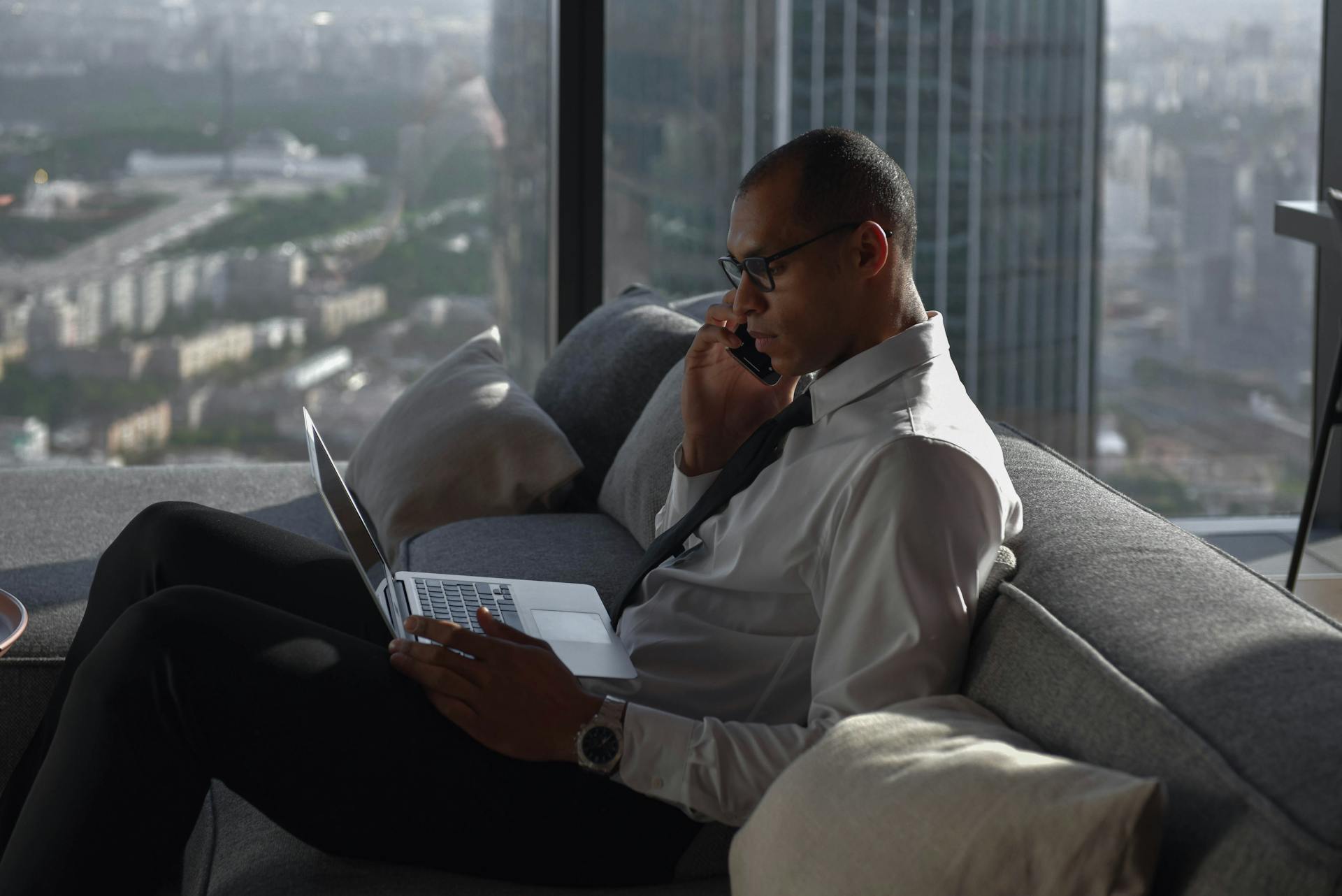 Businessman working on a laptop while on a phone call in a modern office setting with city view.