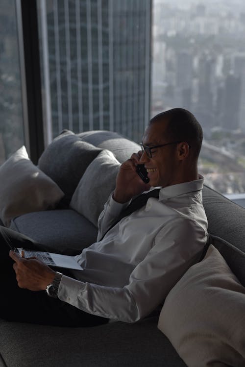 A Man in White Long Sleeves Sitting on the Couch while Talking on the Phone