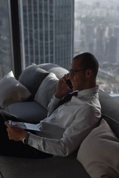 Smiling businessman talking on phone and using laptop indoors with city view.