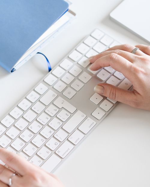 Free Close-up of hands typing on a sleek white computer keyboard with a blue notebook nearby. Stock Photo