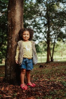 A cheerful young girl with curly hair stands by a tree wearing a yellow shirt and denim shorts outdoors.
