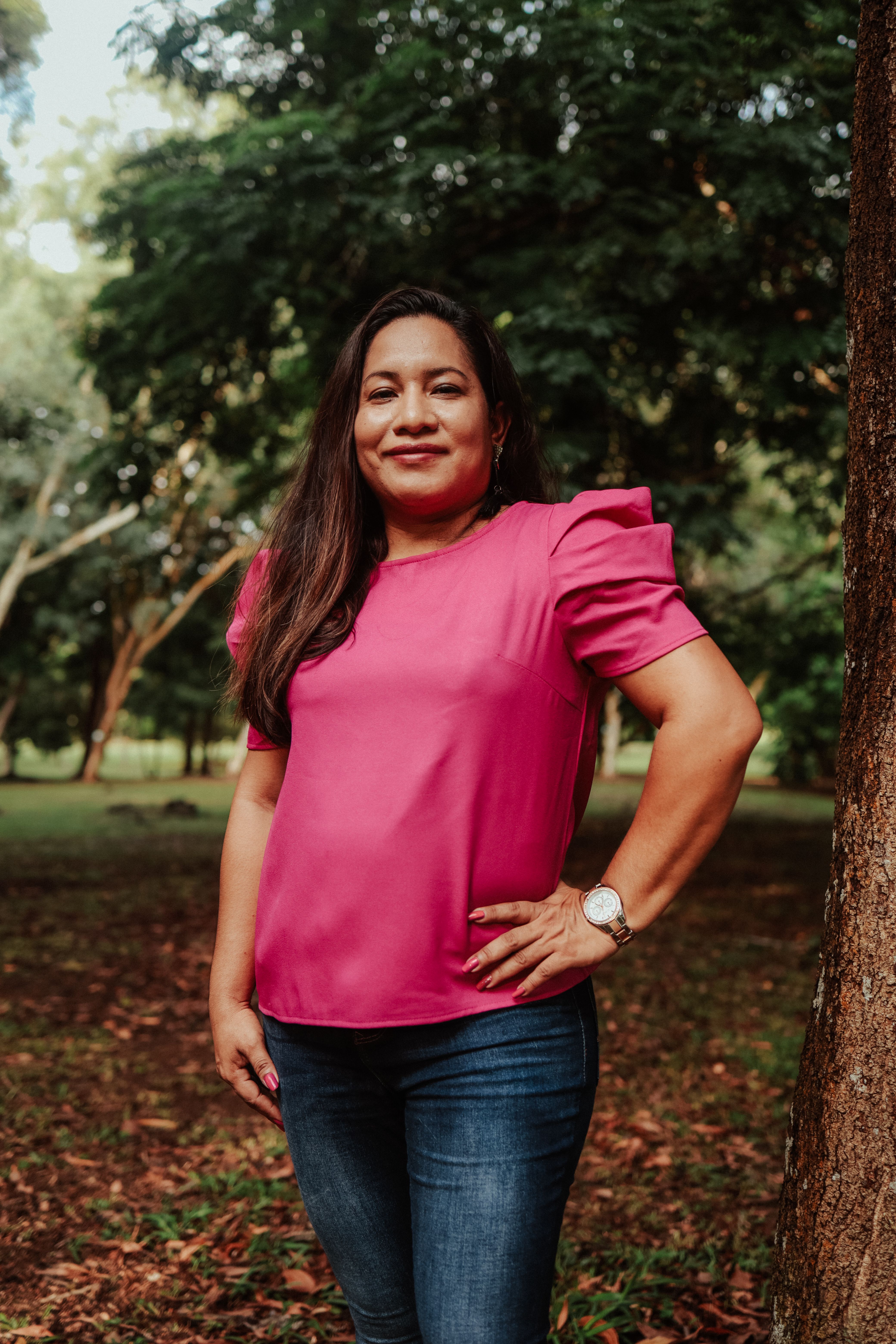 Smiling woman in a park wearing a pink shirt, exuding confidence with hand on hip.