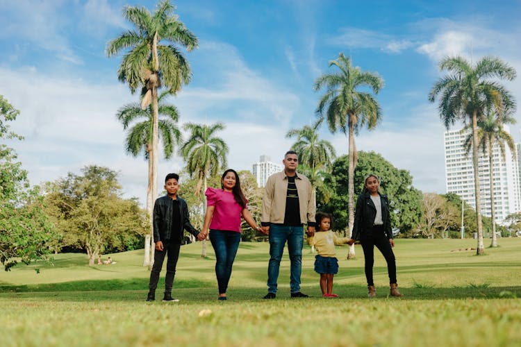 Parents With Three Kids Standing On The Grass Field Holding Hands