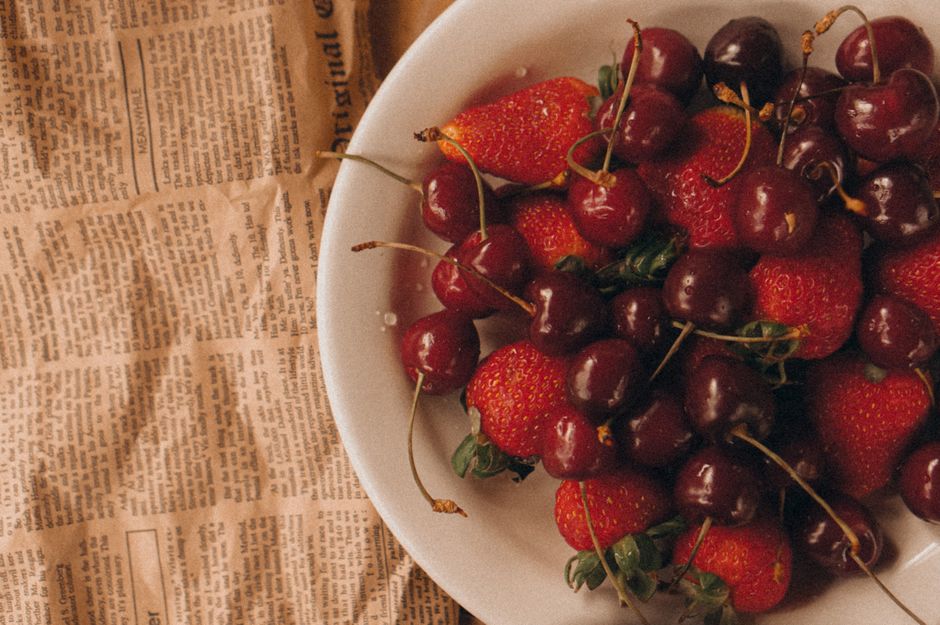 A white plate of fresh cherries and strawberries sits on a vintage newspaper, creating a rustic and summery vibe.